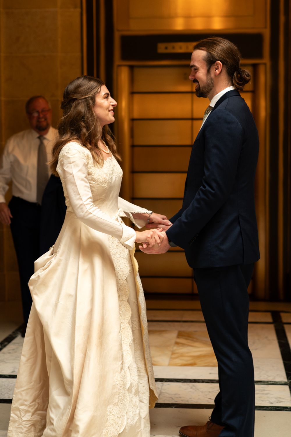 Bride in elegant long-sleeve white gown holds hands with groom in navy suit, smiling joyfully in a warmly lit venue.
