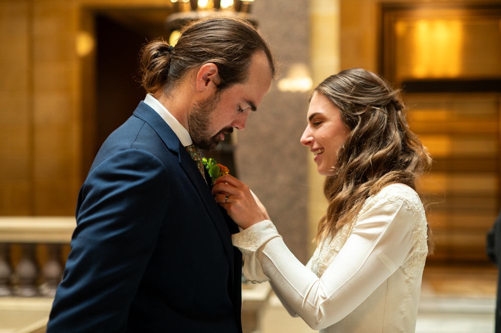 Bride in white dress adjusting groom's boutonniere on blue suit during wedding ceremony indoors.