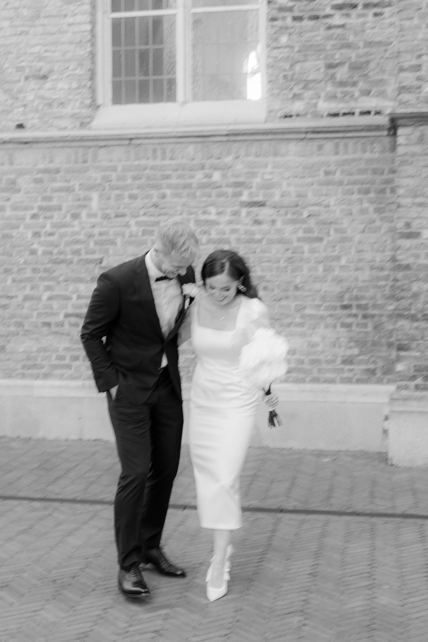 Black and white photo of a bride and groom walking together outside a brick building on their wedding day.
