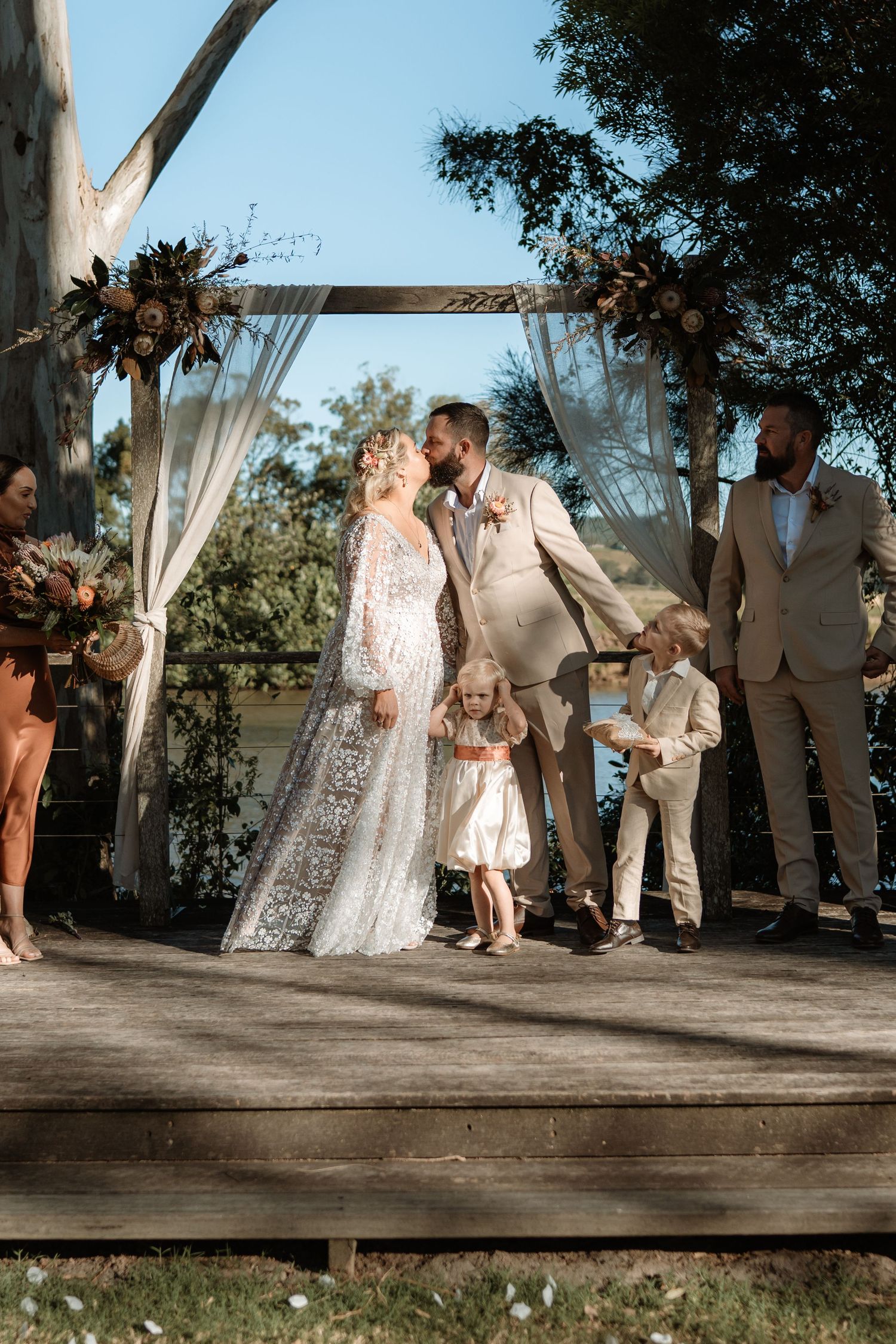 Bride and groom share first kiss at outdoor boho wedding ceremony on wooden deck with floral arch and children nearby.