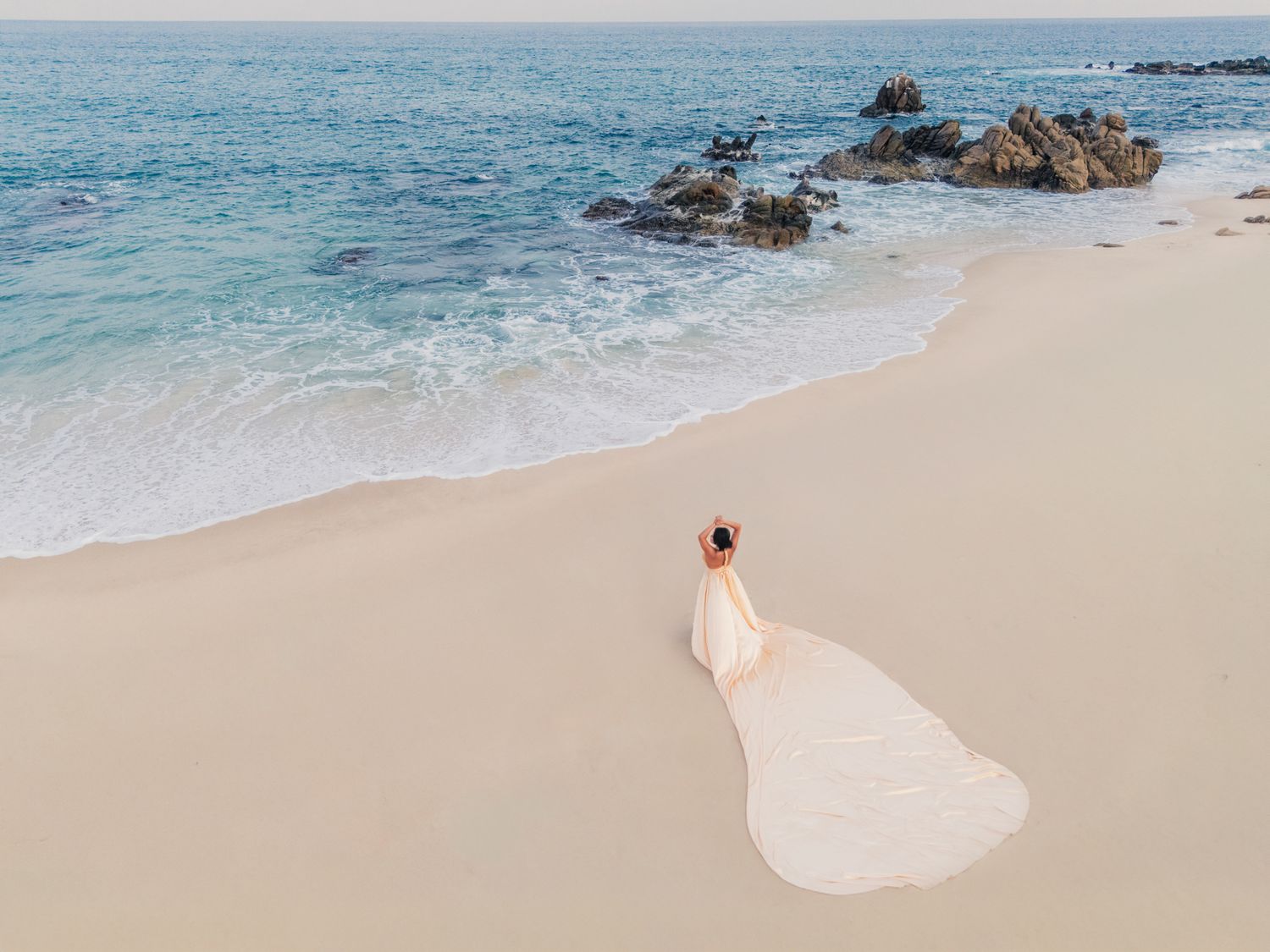 Aerial view of a woman in a flowing purple dress standing on a white sandy beach beside turquoise ocean waves.