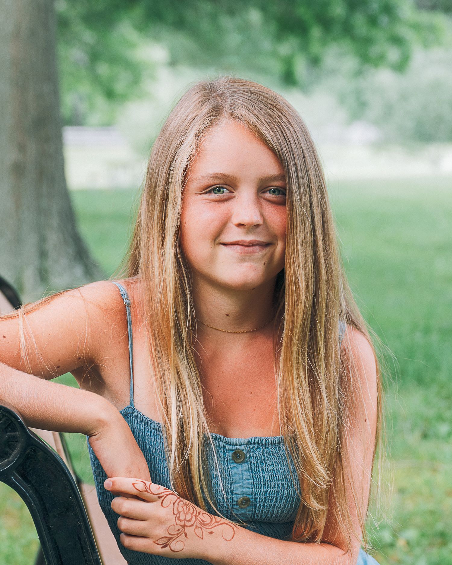 Young girl with long blonde hair smiles outdoors, wearing a denim top with a henna tattoo on her arm.