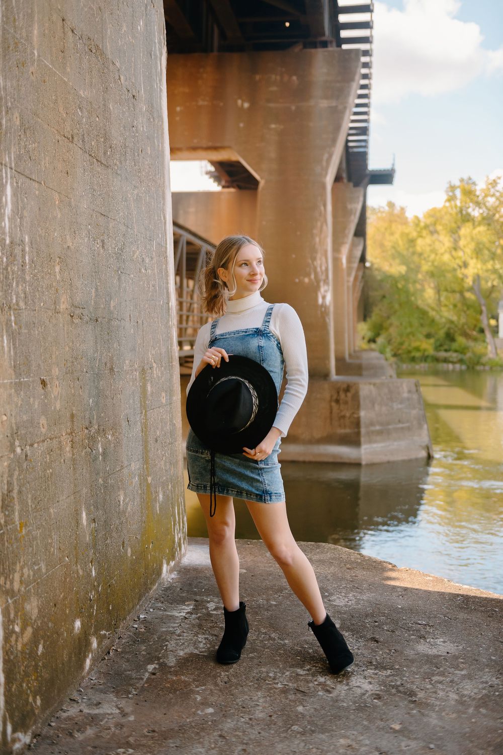 A person in a denim dress and sweater poses under a bridge near a river on a sunny autumn day.