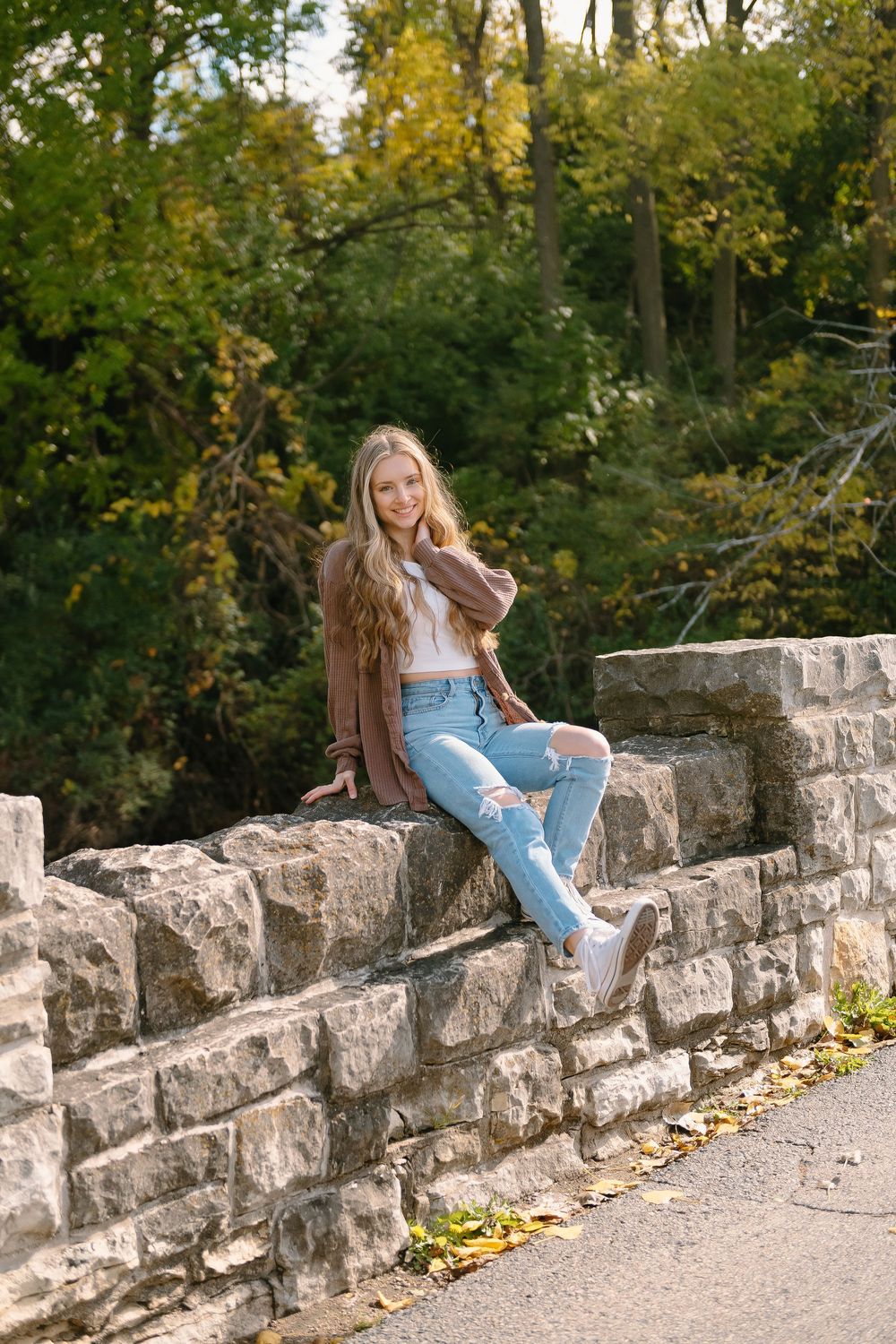 A woman wearing a brown jacket and blue jeans sits on a rustic stone wall surrounded by autumn foliage.