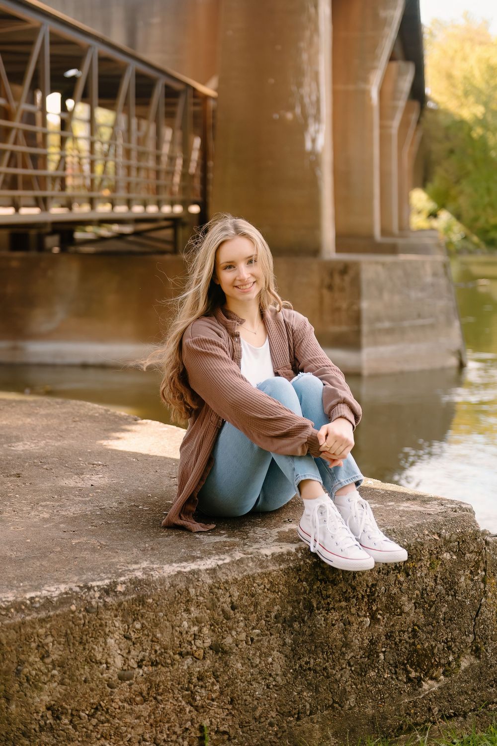Person sitting on concrete ledge near river wearing brown jacket and jeans on autumn afternoon with bridge in background.