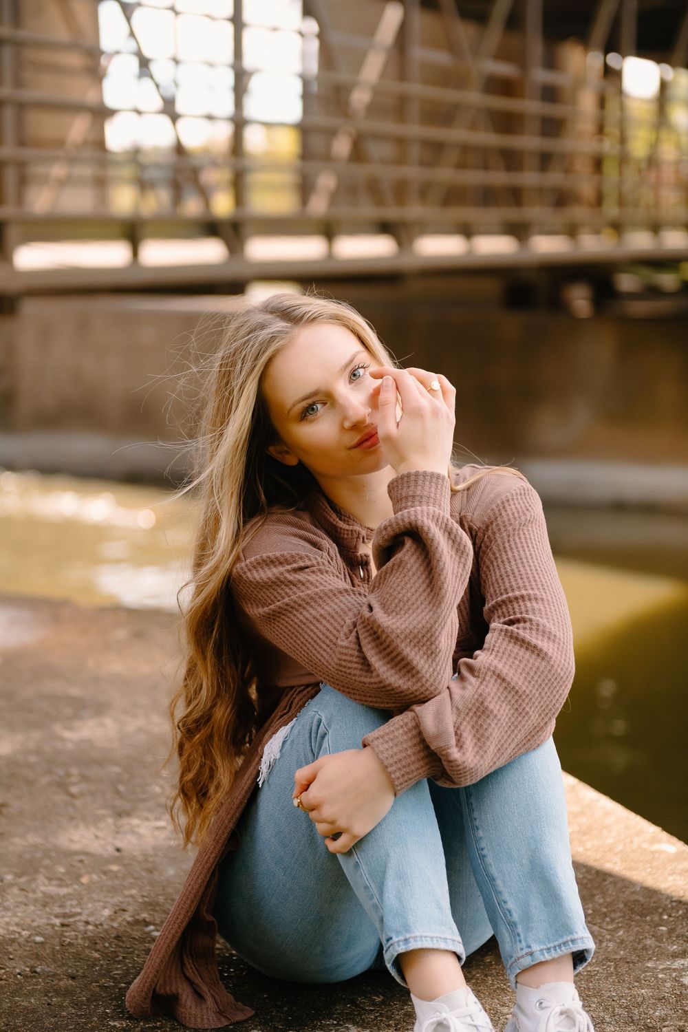 Young graduate woman in brown sweater and light jeans sits casually by water on autumn afternoon.