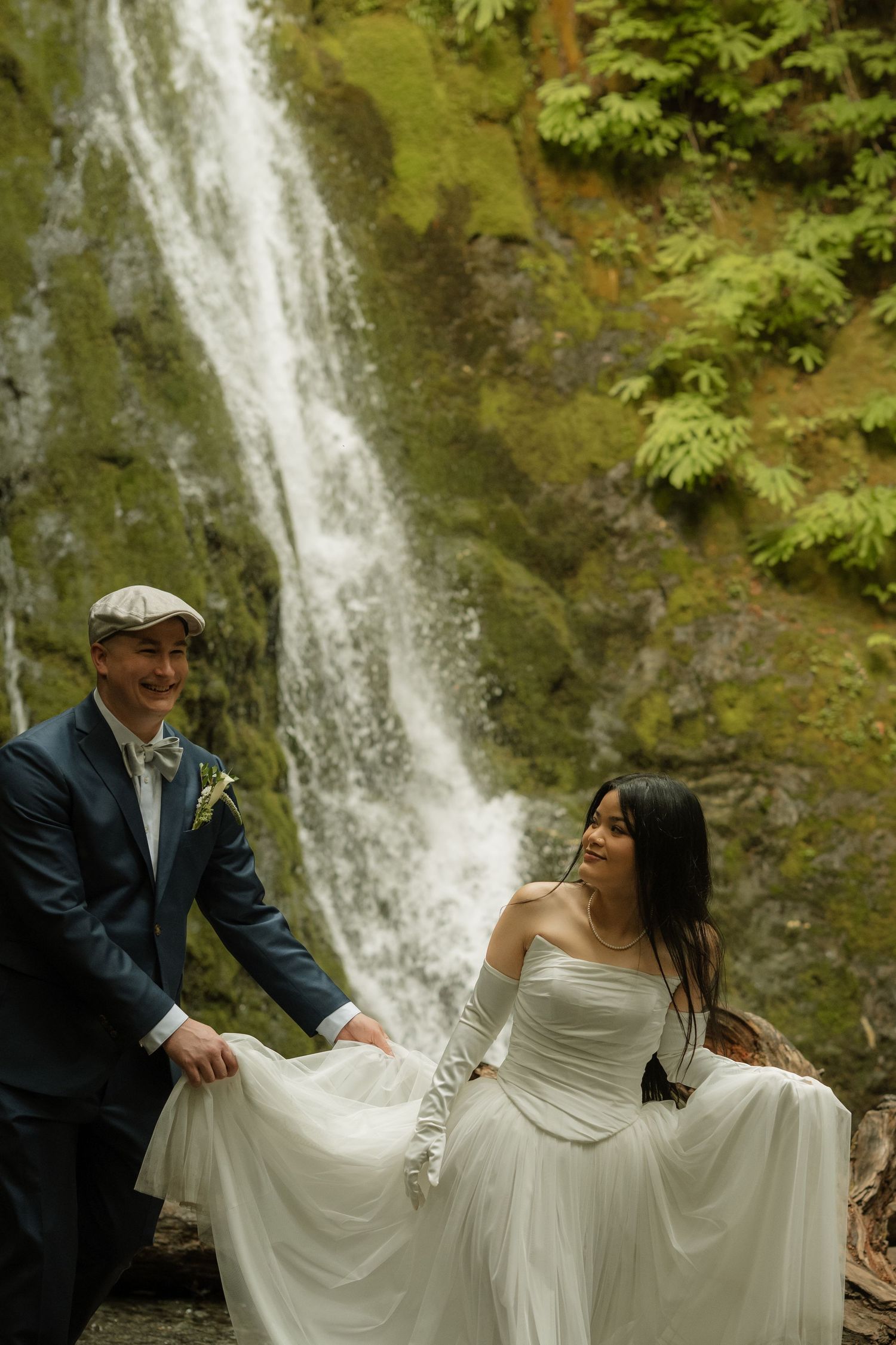 Bride and groom stand before a stunning waterfall on rustic logs in a lush forest wedding elopement.