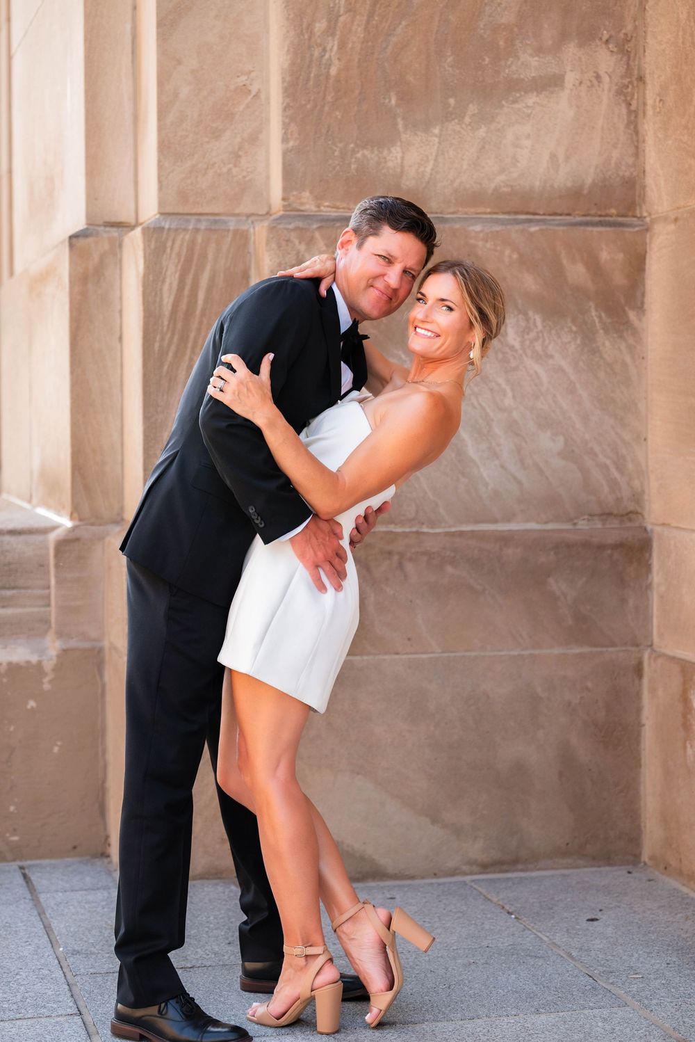 Couple in wedding attire embracing and smiling against a stone wall, woman in white mini dress and man in black tuxedo.