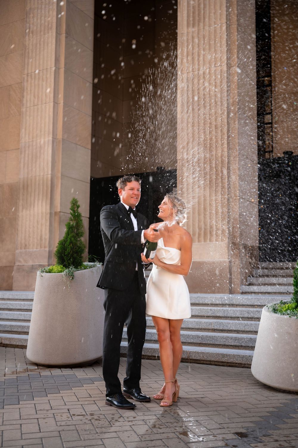 A joyful couple in wedding attire celebrates with champagne spray on elegant building steps.