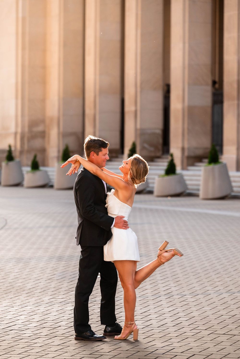 Couple dancing elegantly outdoors, woman in white mini dress and heels, man in black suit, near grand columns.