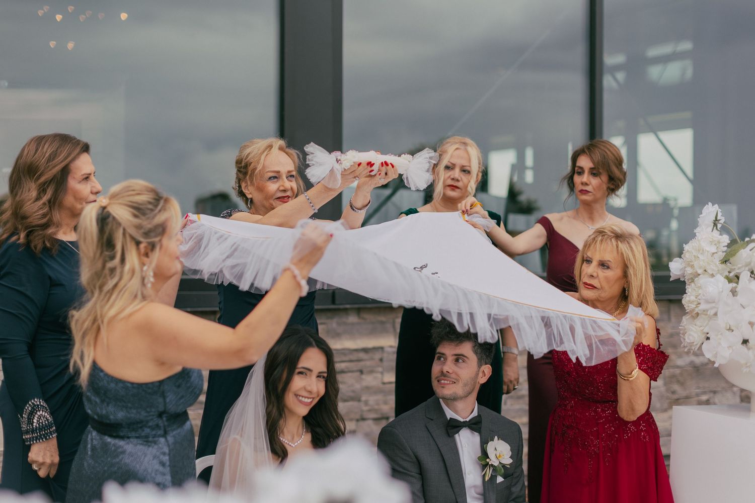 Wedding guests holding a white fabric over bride and groom during traditional ceremony indoors.