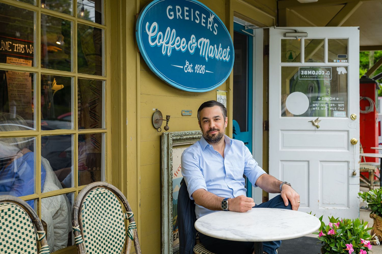 Man sitting outside Greiser's Coffee & Market cafe at a small round table, relaxed in a light blue shirt.