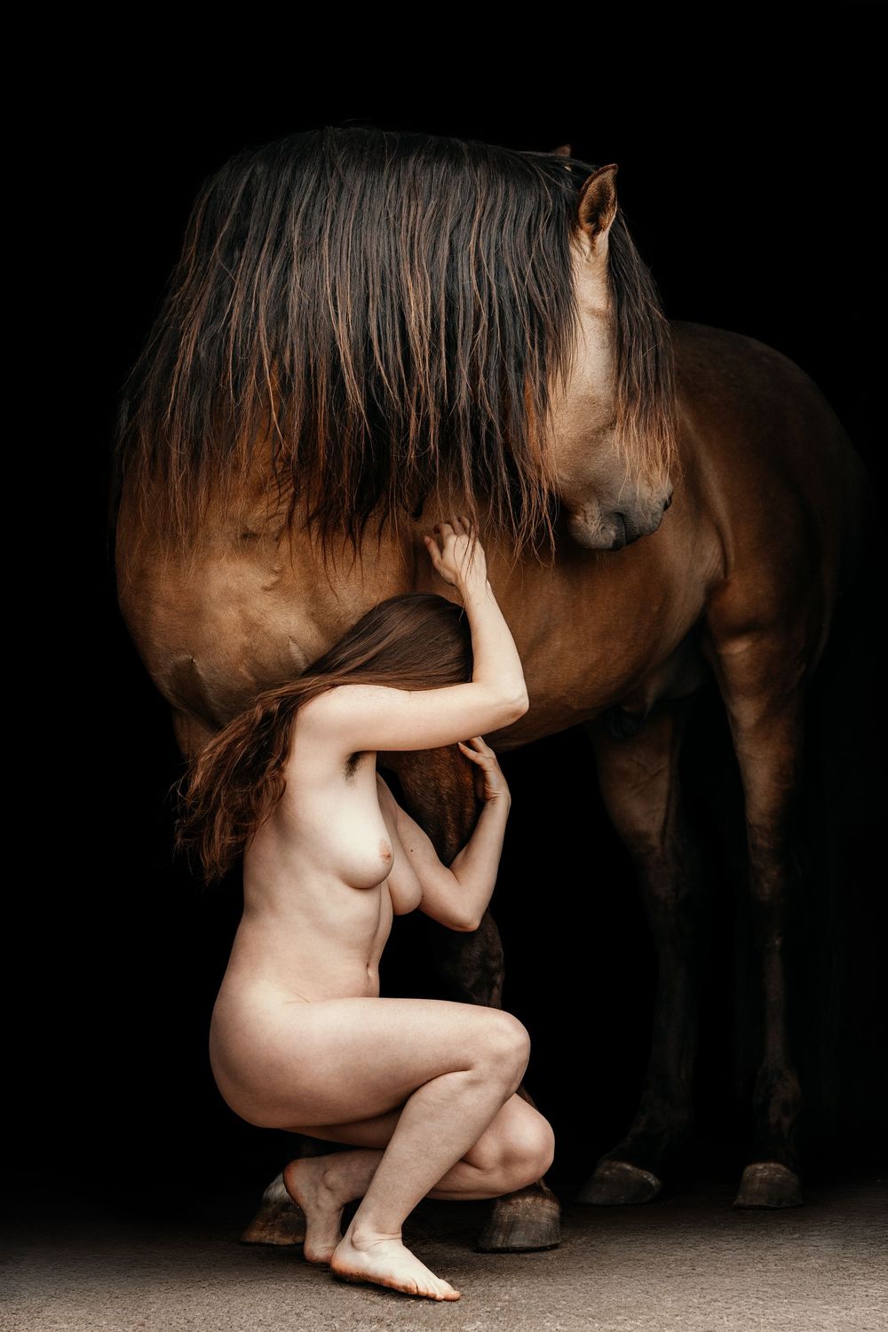 A nude woman crouches beside a brown horse against a dark background in a fine art photography composition.