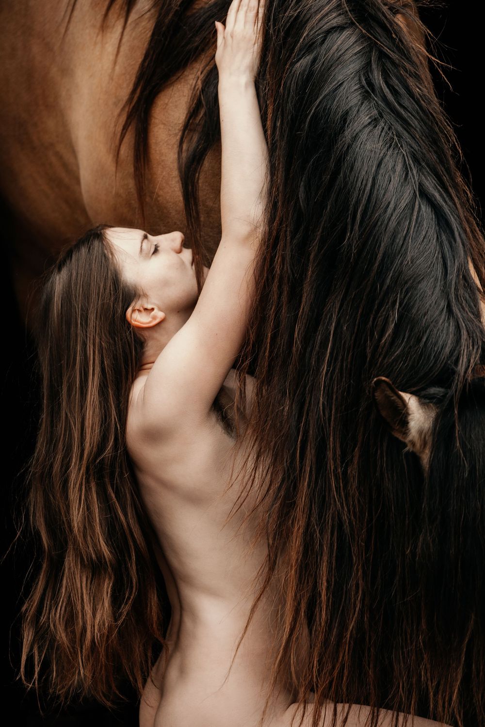 Young woman with long hair reaching up to touch a dark horse's mane against a black background.