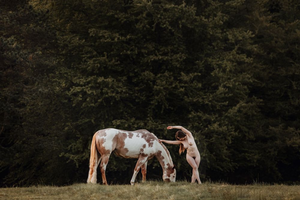 A nude woman stands beside a brown and white paint horse grazing in a meadow with dark green trees behind.