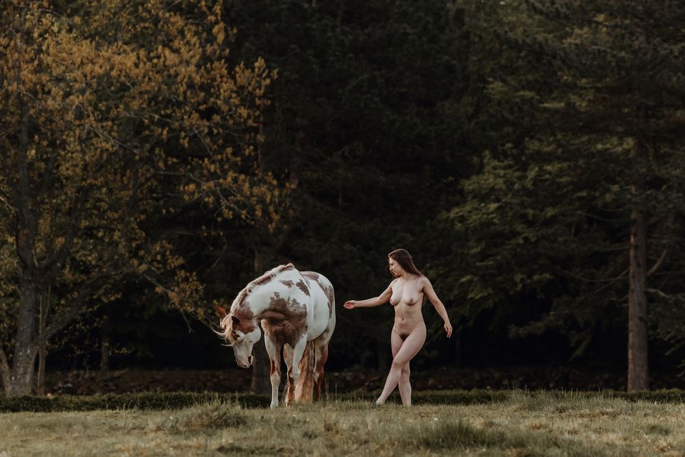 A nude woman stands in a forest meadow, gently touching a white and brown horse among autumn trees.