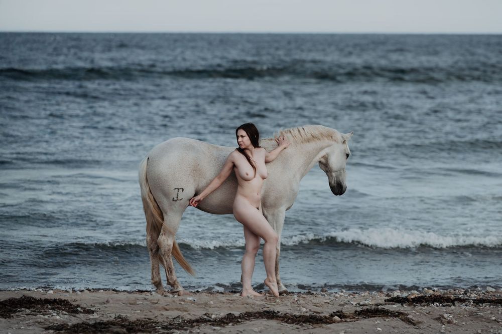 Nude woman standing beside a white horse on a rocky beach with ocean waves in the background.