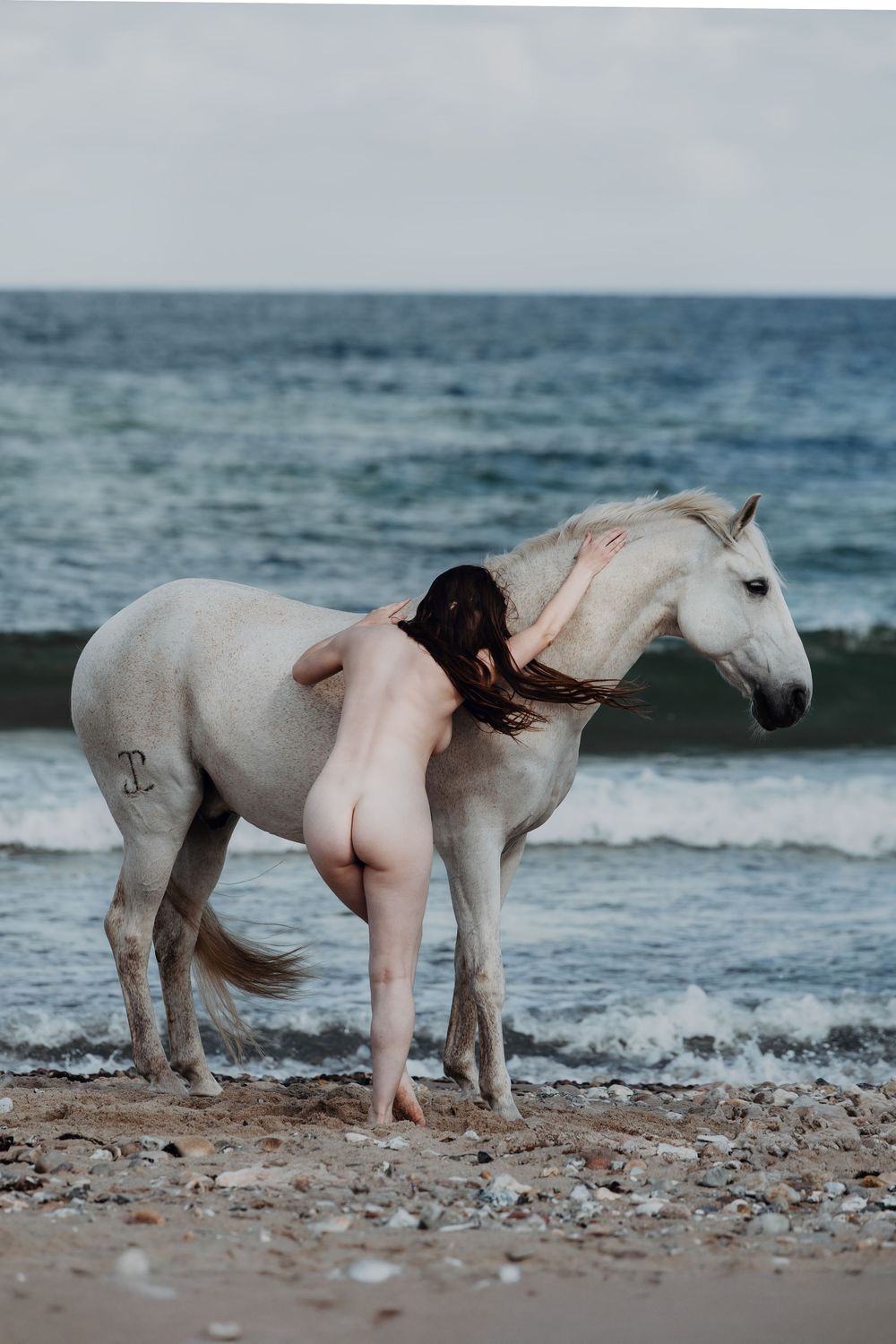 Nude woman embracing a white horse on a rocky beach with ocean waves in the background.