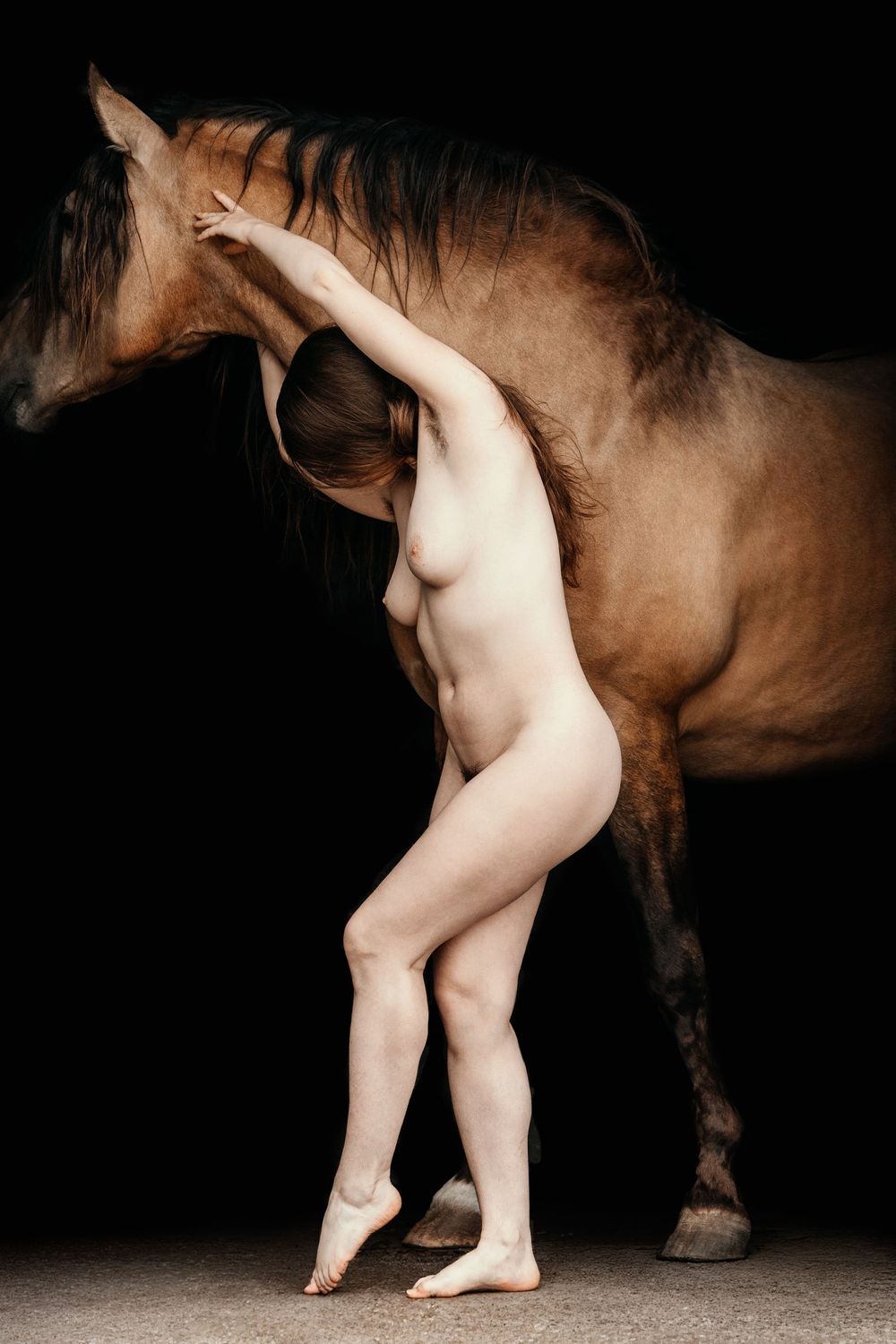 A nude woman leans gracefully against a brown horse against a dark background in a fine art photography composition.