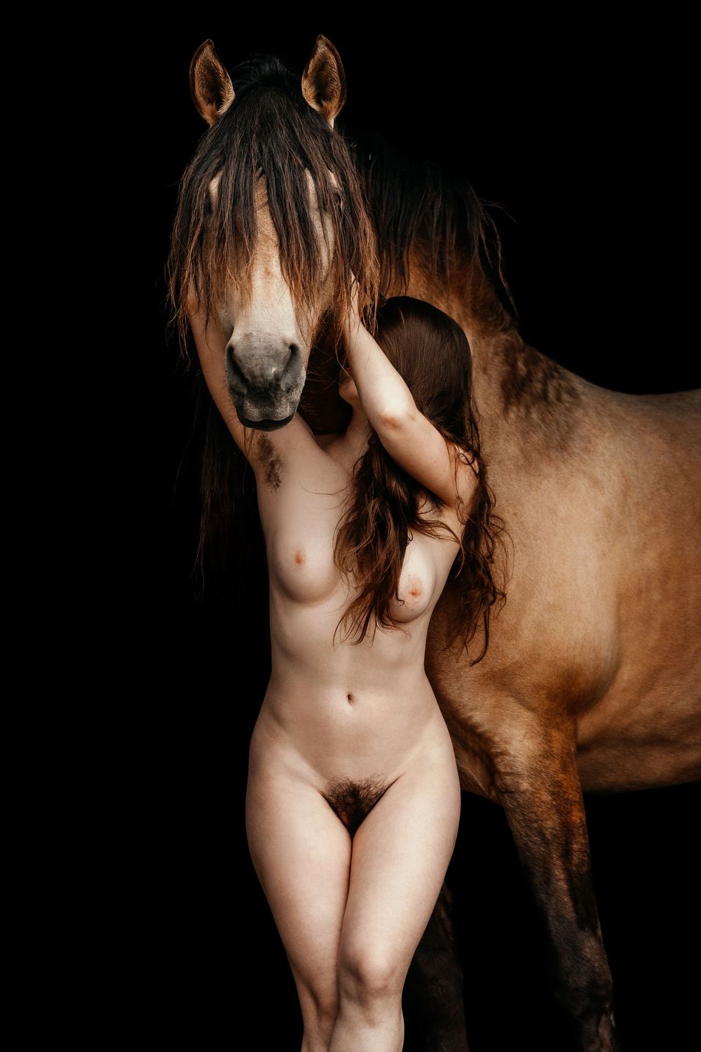 Nude woman standing beside a brown horse against a dark black studio background.