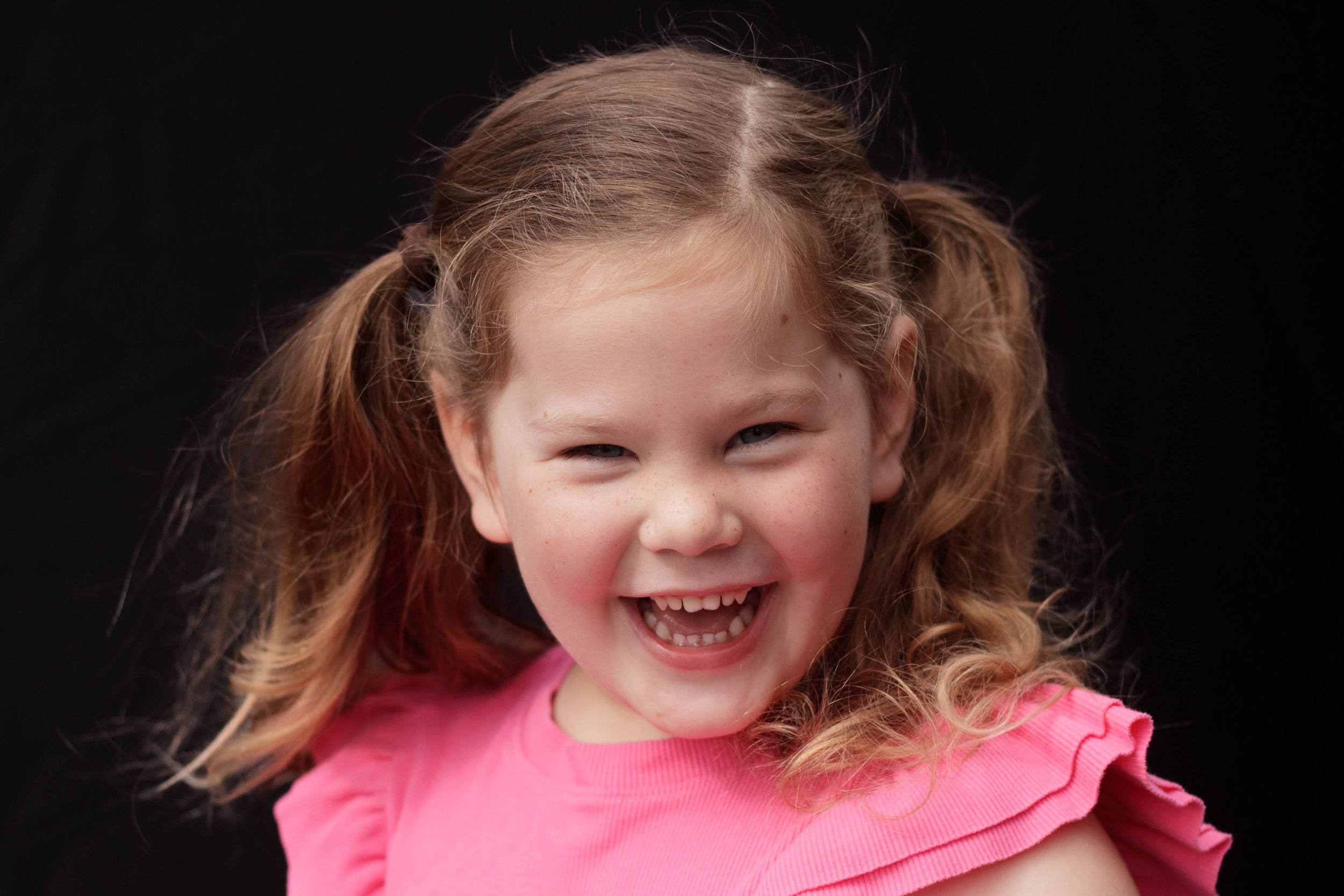 Happy young girl with pigtails laughing in a pink top against a black background.
