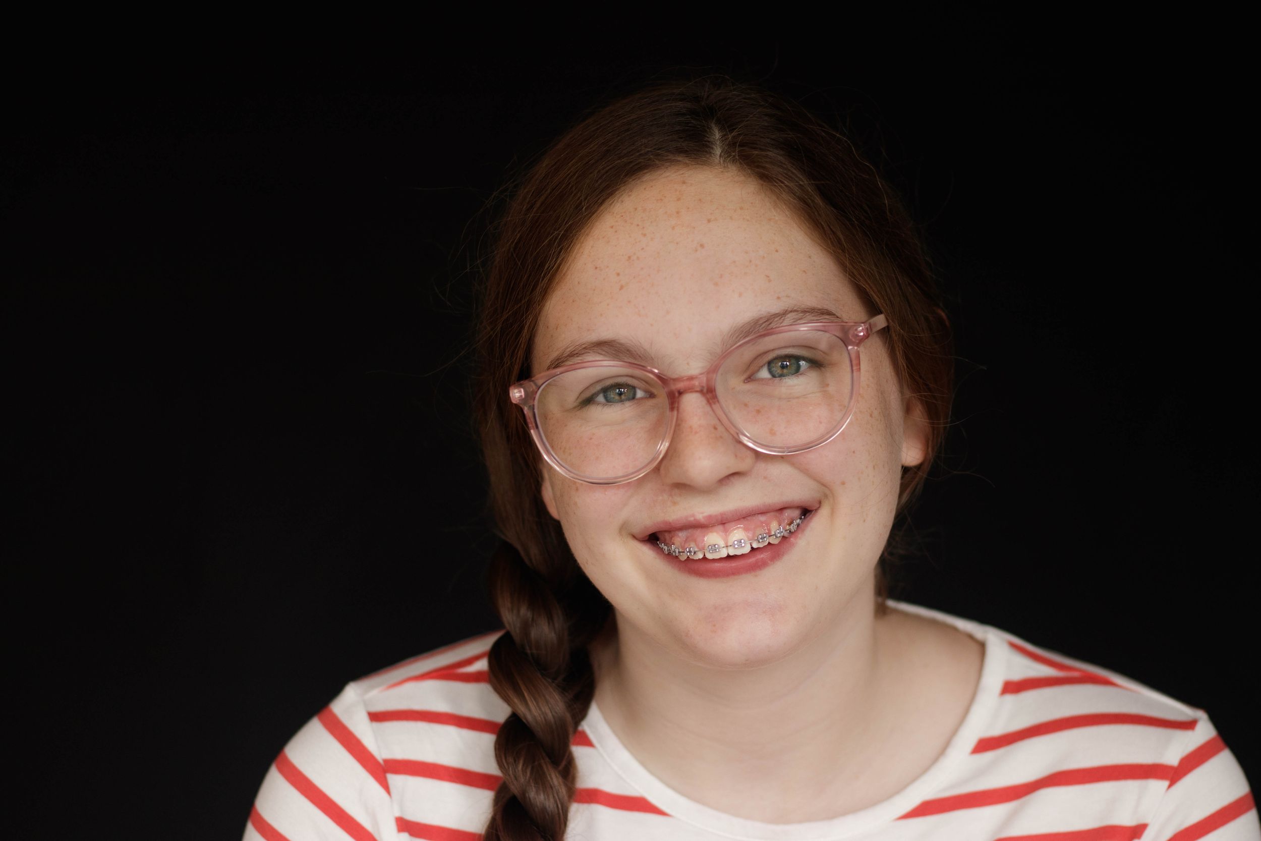 Smiling teenage girl with braided hair, glasses, and braces in a red striped shirt.