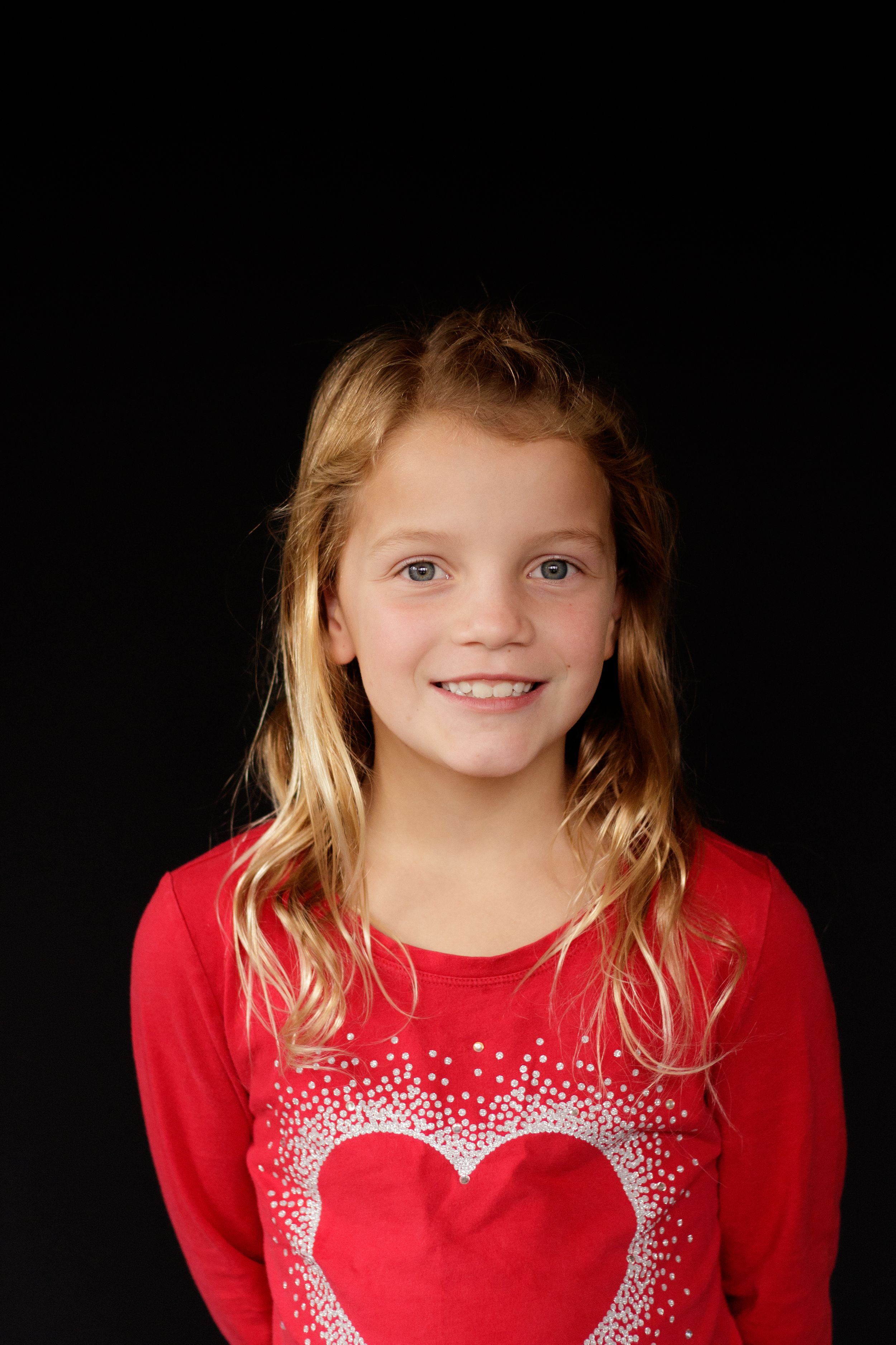 Young girl with long blonde hair smiling in a red heart-print shirt on black background.
