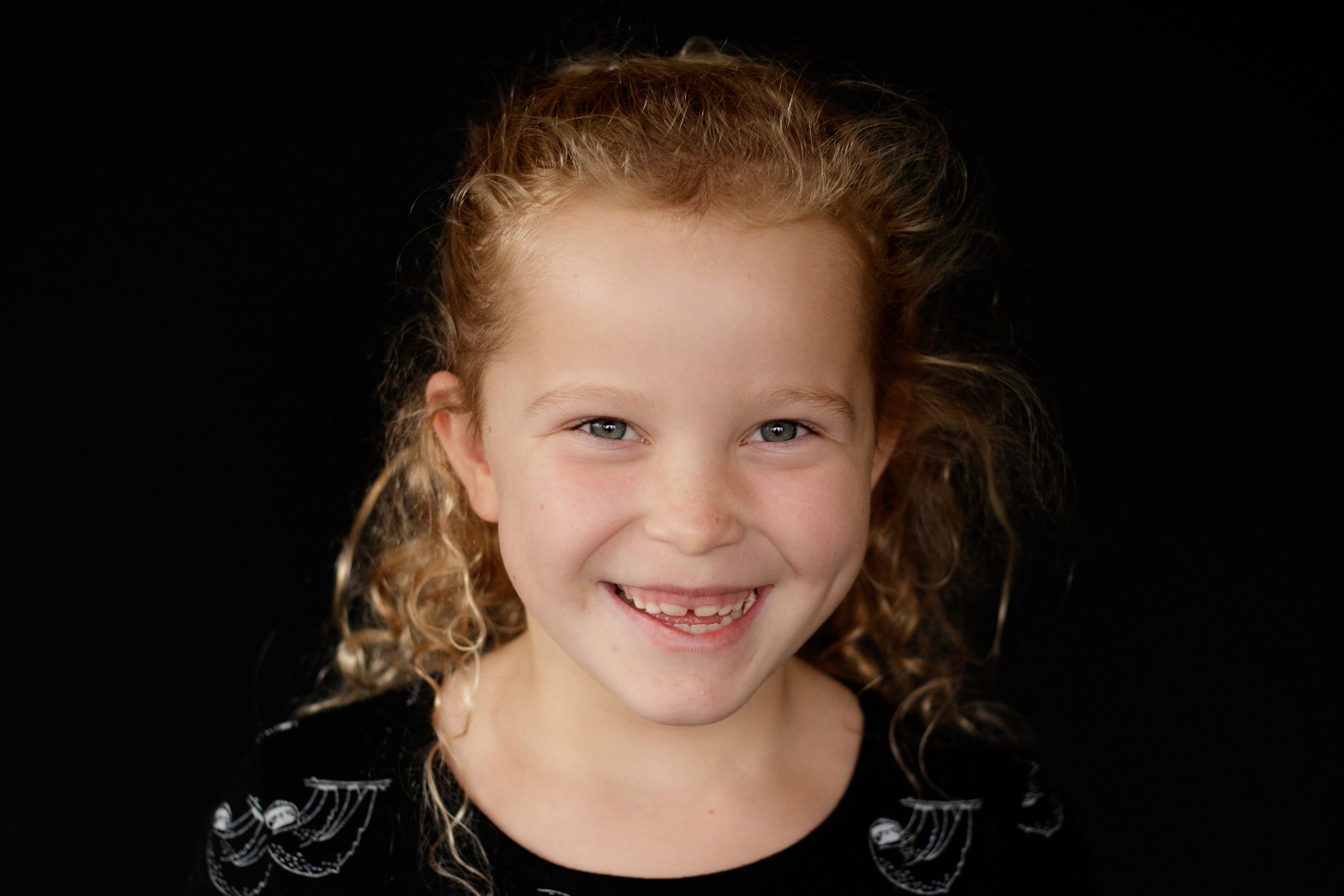 Young curly-haired girl smiling broadly in a black sparkly top against dark background.