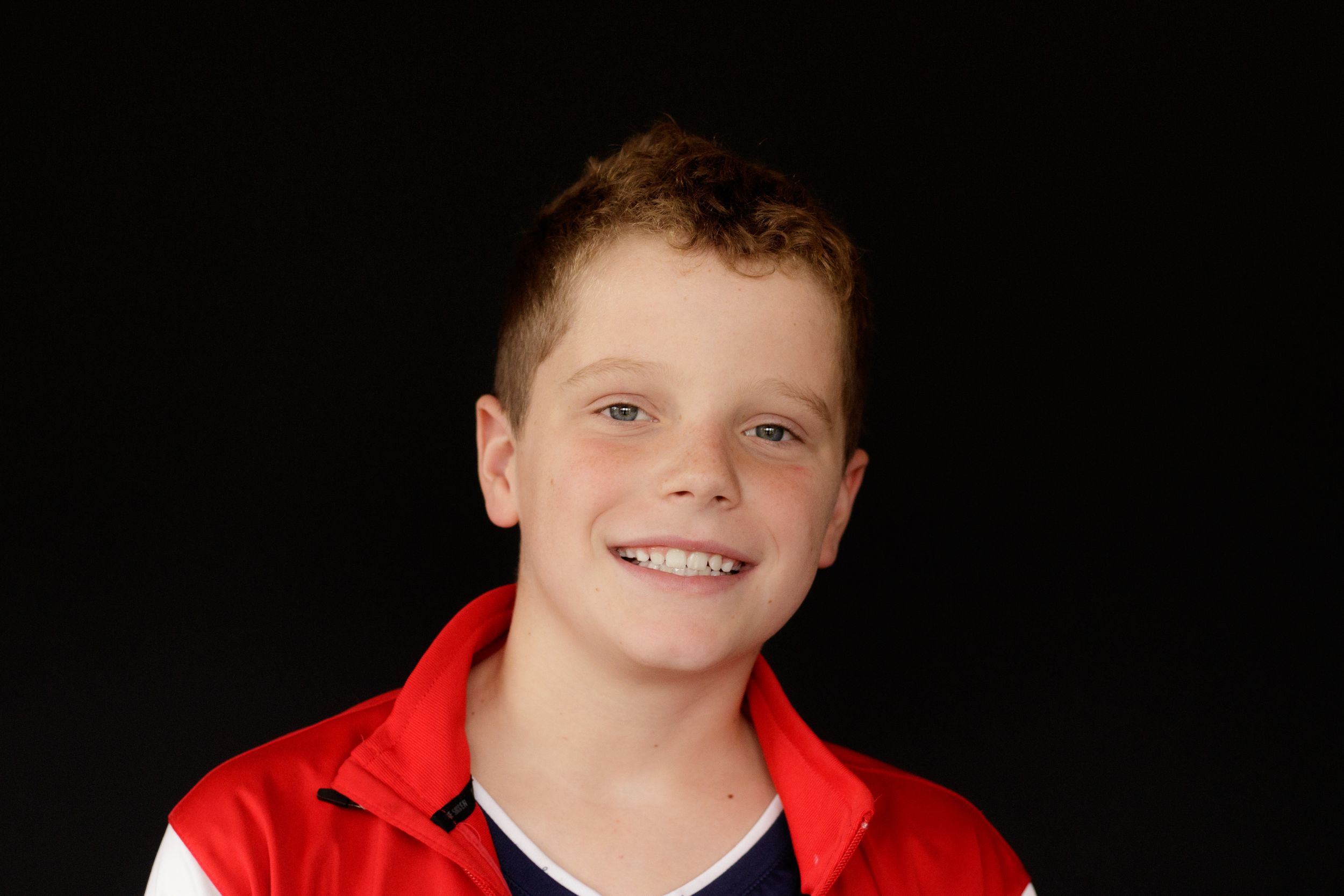 Smiling young boy with red hair wearing a red and blue jacket against a dark background.