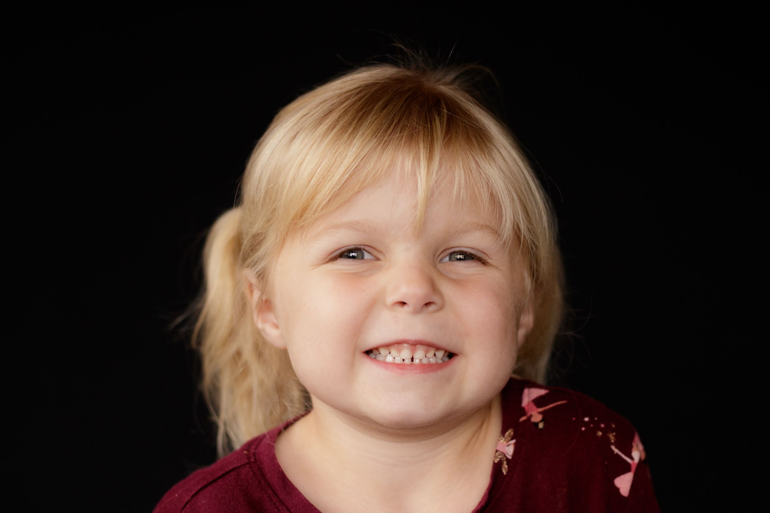 Adorable blonde toddler girl smiling cheerfully in a dark maroon floral top.