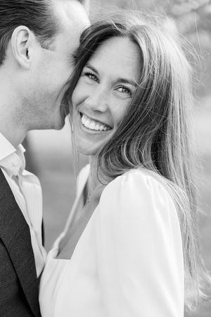 Black and white photo of a smiling woman in white dress with a man in suit whispering in her ear outdoors.