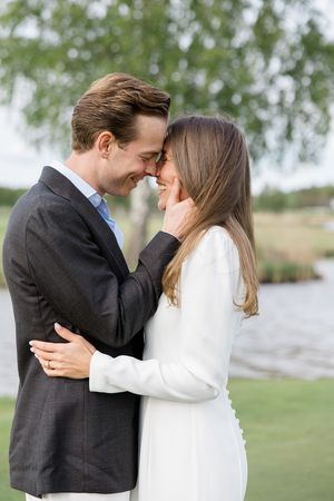 Couple sharing a romantic forehead touch outdoors, man in gray suit, woman in white dress, near a serene lake.