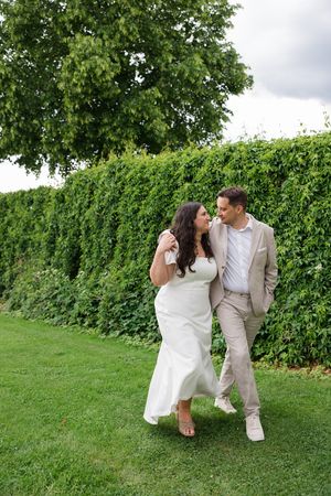 A couple in wedding attire walks and laughs together on green grass beside a lush hedge outdoors.