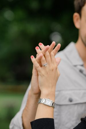 Close up of hands showing engagement ring with silver watch on wrist against blurred natural background.