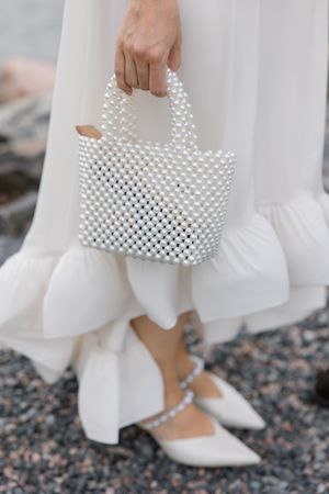 A white beaded handbag is held alongside a flowing white dress and matching white shoes on a textured ground.