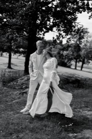 A couple in all-white outfits embraces romantically in a park setting, captured in black and white photography.