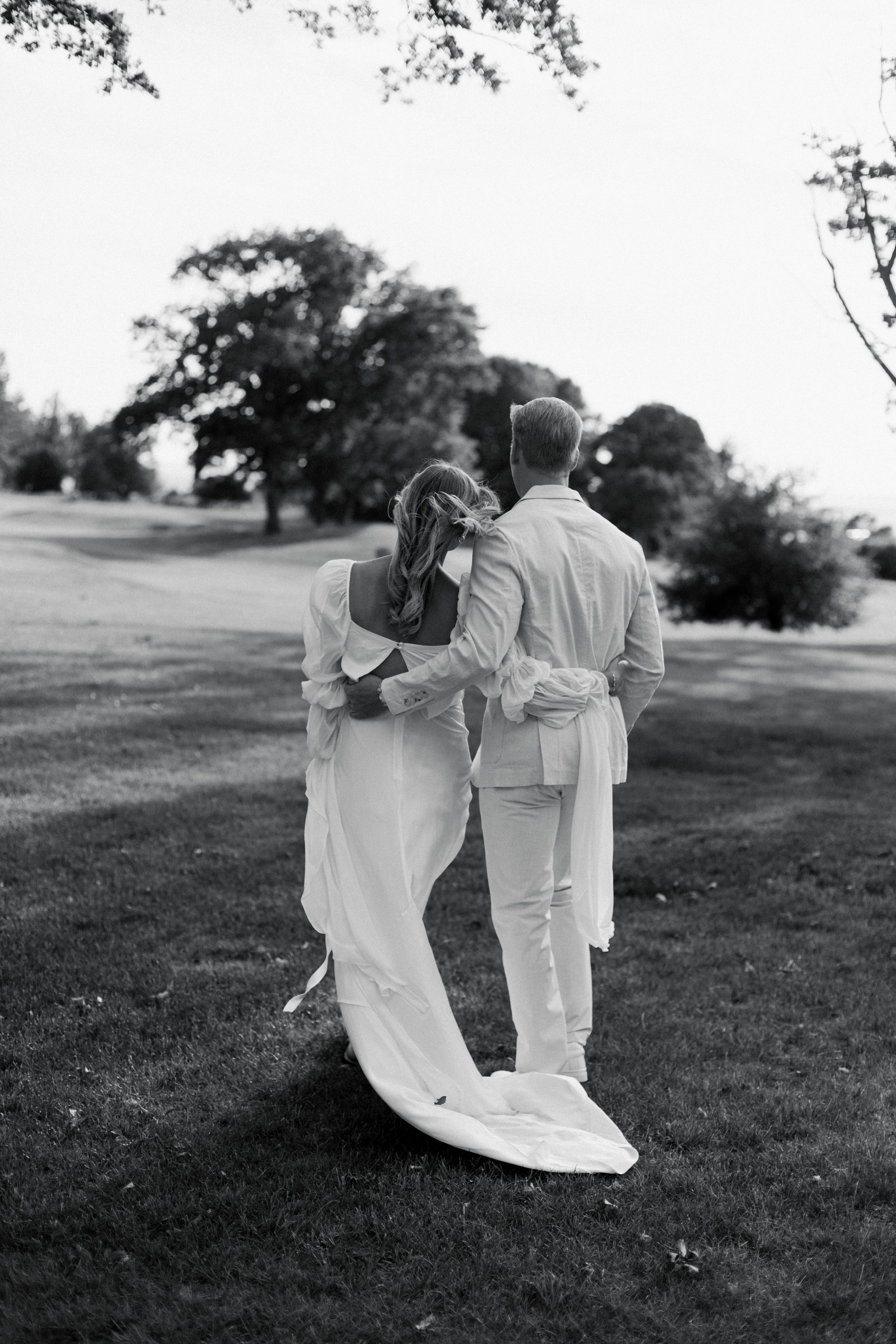 A couple in white attire embraces while walking on a grassy hill with trees in the background, captured in black and white.