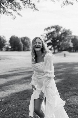 Black and white photo of someone wearing a flowing white dress laughing joyfully in a sunny park setting.