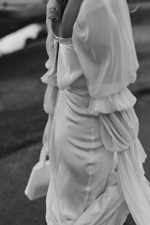 Black and white detail shot of a bride wearing a flowing white dress with gathered fabric and holding a small purse.