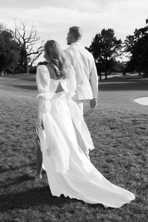 A couple in white wedding attire stand together on a grassy lawn at dusk with the bride's dress flowing in the breeze.