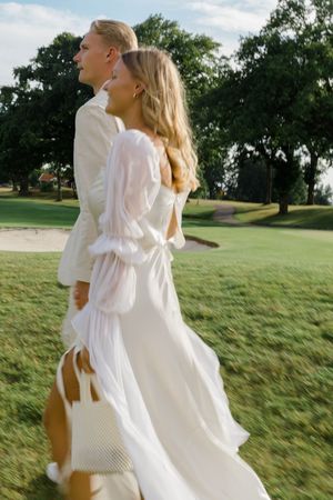 A couple in white attire walking together on a grassy golf course on a sunny day.