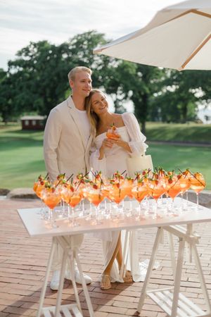 A couple in white stands beside a table of orange signature cocktails on a deck overlooking a golf course.