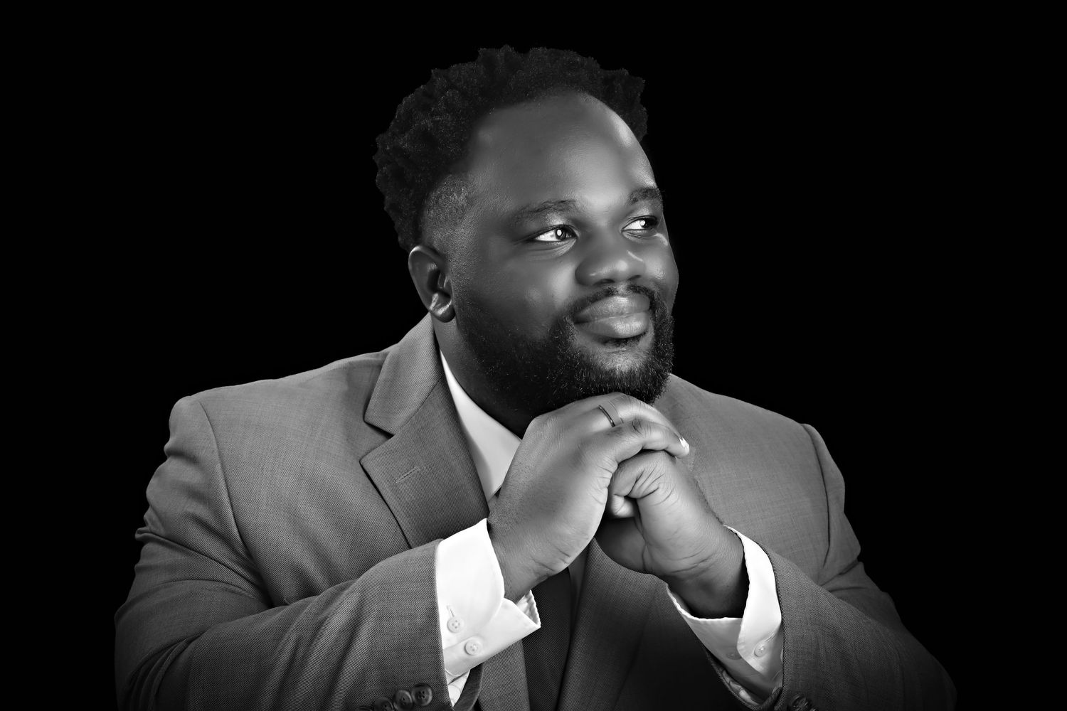 Black and white portrait of a man in a suit with clasped hands, looking upward thoughtfully.