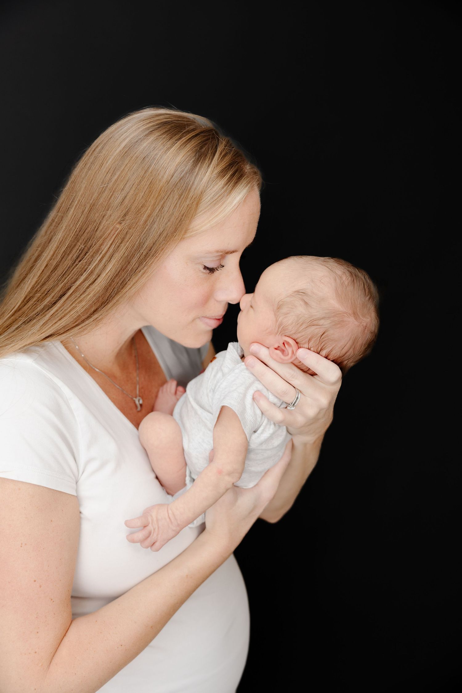 A tender moment as a blonde mother in white gently holds and nuzzles her sleeping newborn baby against a black background.