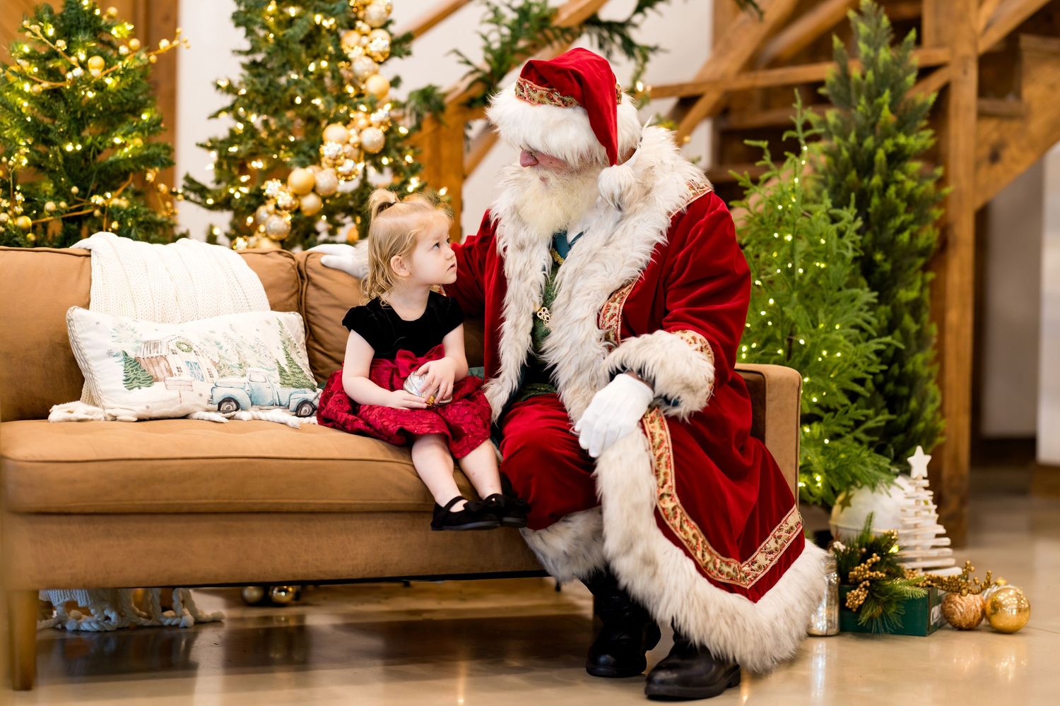 Young girl in red dress sits with Santa Claus on a couch surrounded by Christmas trees and festive decor.