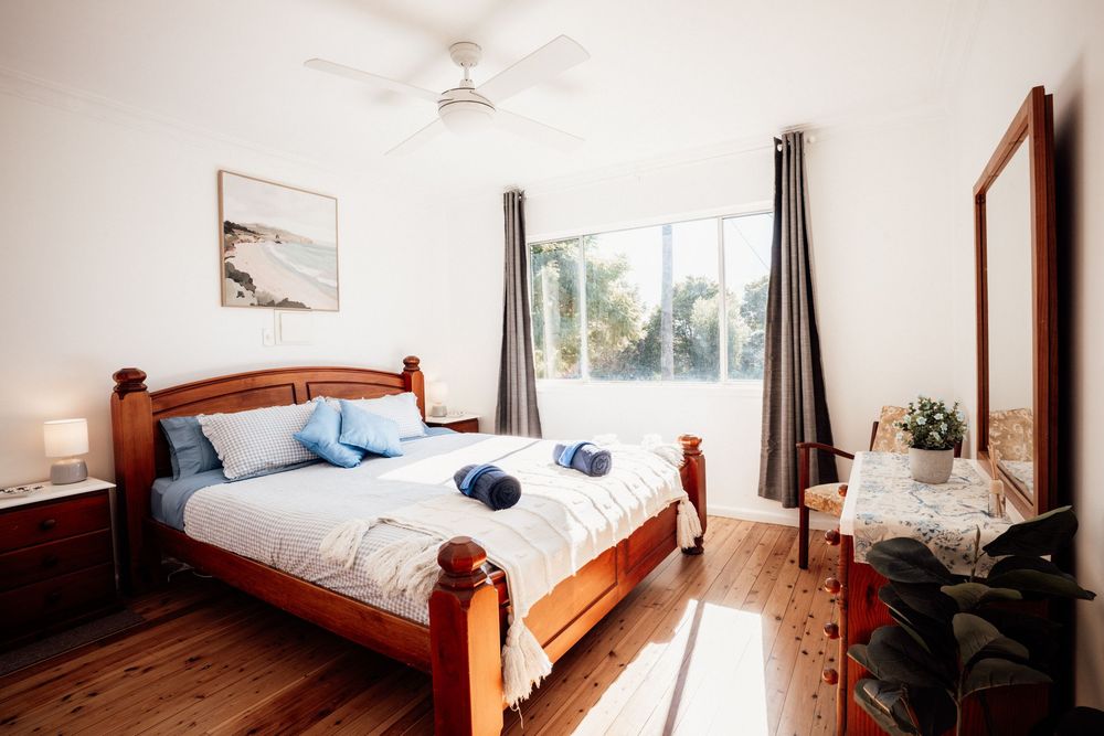 Bright bedroom with timber bed frame, blue pillows, hardwood floors, ceiling fan, and large window with garden views.