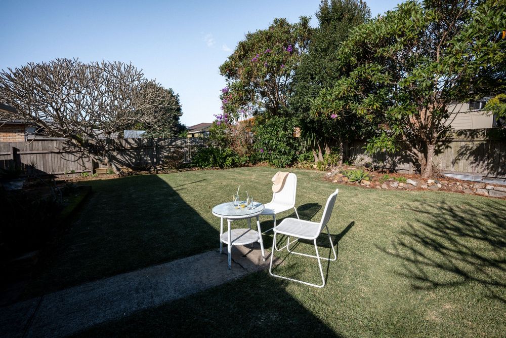 White table and chairs on lush green lawn surrounded by mature trees in a sunny backyard garden setting.