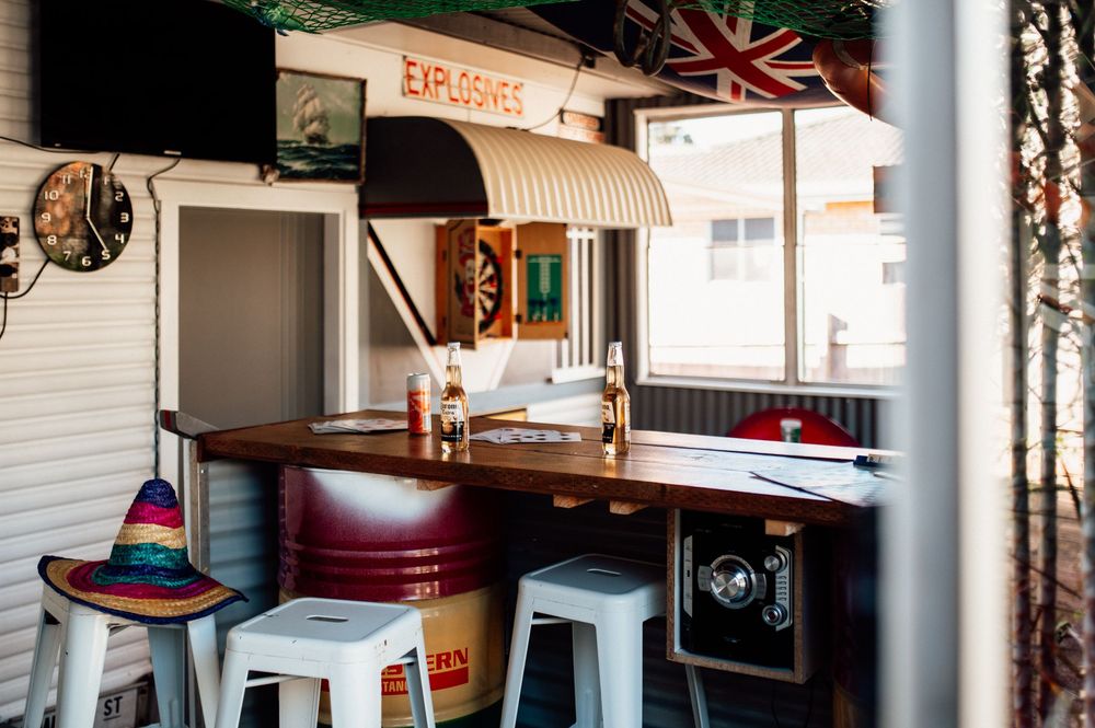 Cozy outdoor bar area with white stools, wooden counter, colorful sombrero, and Explosives sign in background.