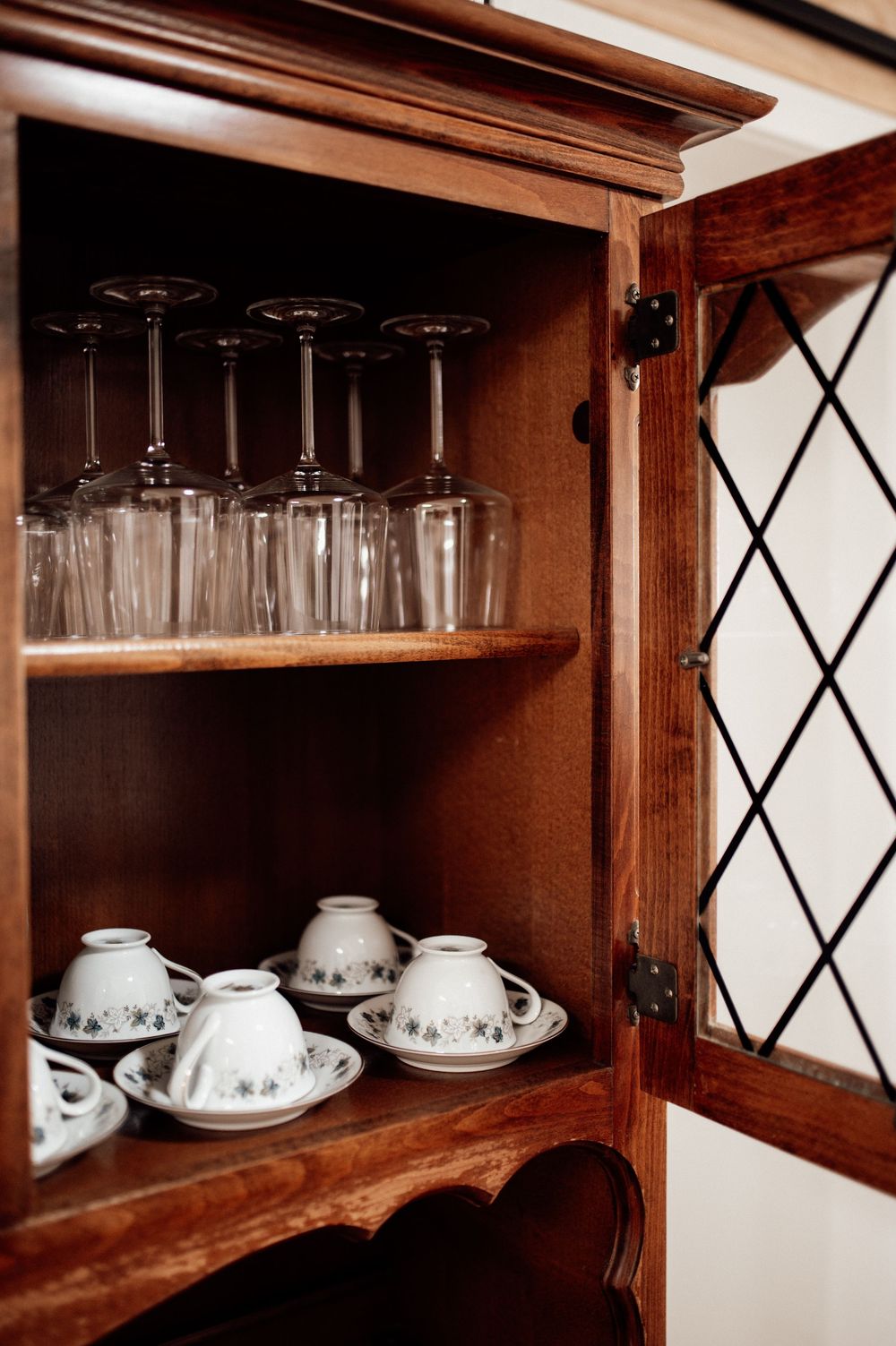 Wooden cabinet with glass-door displaying upside-down wine glasses on top shelf and white floral tea cups with saucers below.