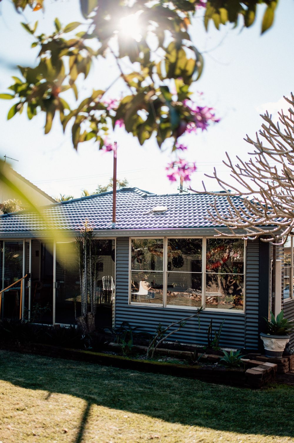 A charming grey weatherboard house with blue roof tiles surrounded by lush garden trees.